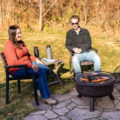 People sitting by Bronze 30 Inch Outdoor Wood Burning Fire Pit With Grill Grate, showing sausages cooking on a sunny day.