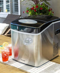 Portable Countertop Ice Maker Delivers 12 Bullet Ice Cubes in 7 Minutes; stainless steel unit on outdoor table, next to drinks.