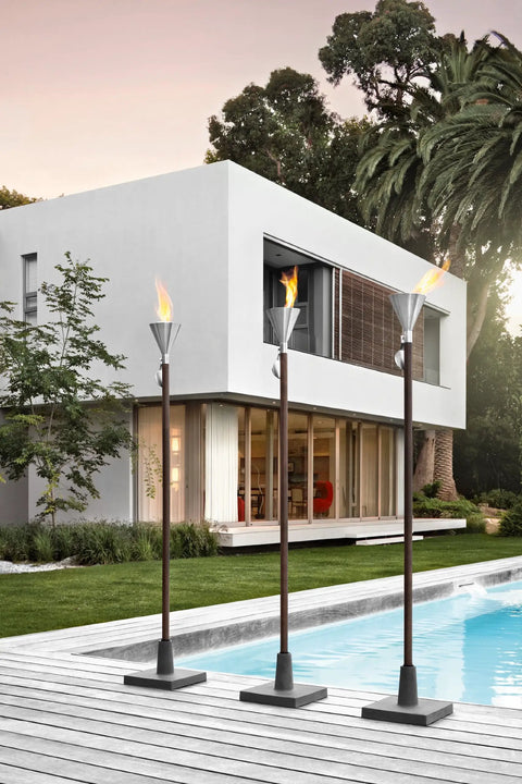 Three flaming torches with black weighted polystone bases on a patio next to a modern house, showcasing outdoor decor.