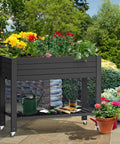 Elevated Garden Bed With Wheels And Storage Shelf For Outdoor Use Plastic Planter Box With Drain Plug Central Divider And Water Level Float System For Precise Moisture Control For Plants Vegetables Flowers And Herbs Black on a patio with flowers and gardening tools.