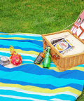 Picnic setup on grass with a colorful blanket, wicker basket, drinks, fruit, and snacks.