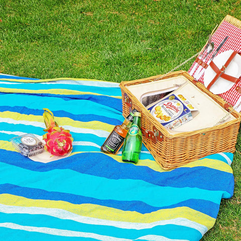 Picnic setup on grass with a colorful blanket, wicker basket, drinks, fruit, and snacks.