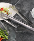 Restaurant Grade 6.5 Inch Pom Tong Set Of 6 Stainless Steel Garnish Tongs with Satin Finish beside sliced vegetables on plates.