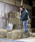 Person using Long Knob Manure Fork With 48 Inch Ash Handle And 13 Inch Head, Forged Carbon Steel Tines in a barn with hay bales.
