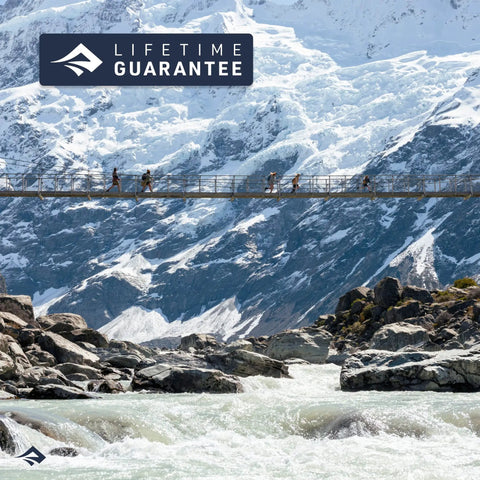 People crossing a suspension bridge over a river with snowy mountains and "Lifetime Guarantee" label visible in the background.