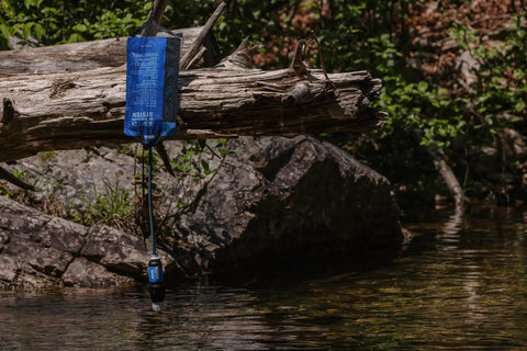 Portable Squeeze Water Filtration System With Two 32 Oz Pouches hanging on branch over water, 0.1 micron inline filter visible.