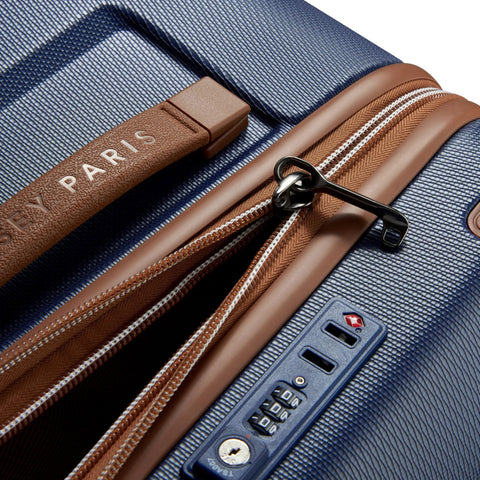 Close-up of Delsey 26-inch spinner luggage with navy blue basketweave texture, brown faux leather accents, TSA lock, and zippers.