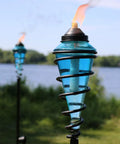 Two Pack Glass Patio Torch Set, Tall Outdoor Torches With Adjustable Height Up To 66 Inches For Garden, Deck, And Backyard, Blue Glass Vessels With Metal Casings, Perfect Ambient Lighting For Evenings, Entertaining, And Ambience, lit by a lakeshore.