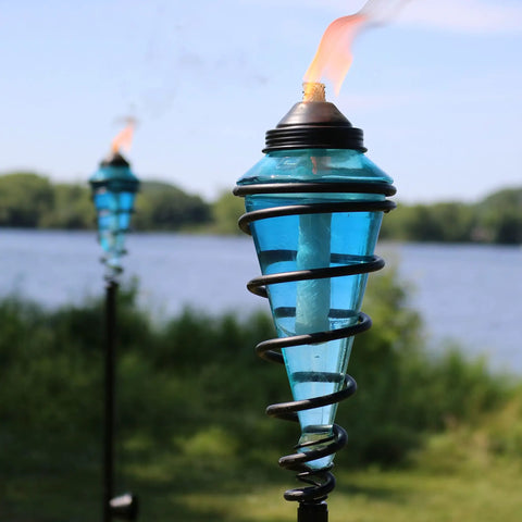 Two Pack Glass Patio Torch Set, Tall Outdoor Torches With Adjustable Height Up To 66 Inches For Garden, Deck, And Backyard, Blue Glass Vessels With Metal Casings, Perfect Ambient Lighting For Evenings, Entertaining, And Ambience, lit by a lakeshore.