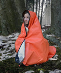 Person in forest wrapped in Emergency Survival Blanket Tarp Two Point Zero Windproof and Waterproof Gear, orange color.