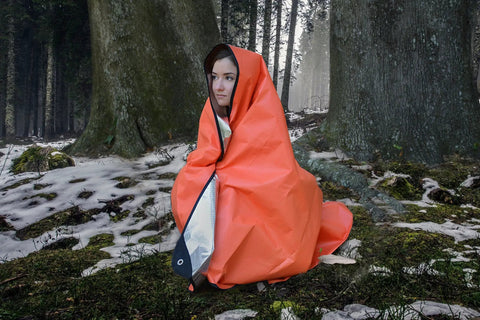 Person in forest wrapped in Emergency Survival Blanket Tarp Two Point Zero Windproof and Waterproof Gear, orange color.