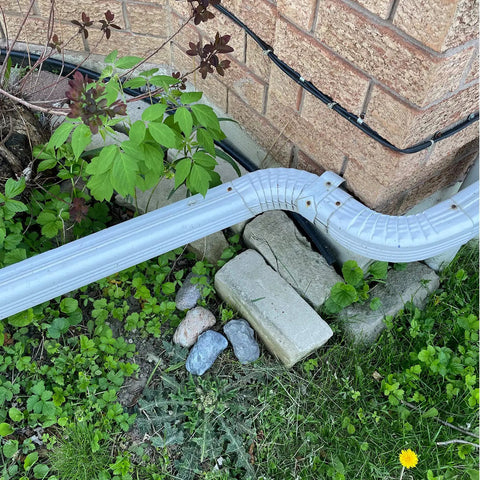 Corner of a brick house with drainage pipe, surrounded by garden plants and stones, hiding stash a spare key in this fake rock that looks like real stone for outdoor gardens and yards geocaching safe key hider type C 1 pack weatherproof discreet outdoor decor.