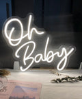 Oh Baby Warm White LED Neon Sign For Home Decor And Party Vibes Great For Bachelorette Parties Birthday Weddings Engagements Bar Pub Club Wall Hanging Neon Light For Event Decor And Social Gatherings In Any Room, glowing on a stand with decor items.