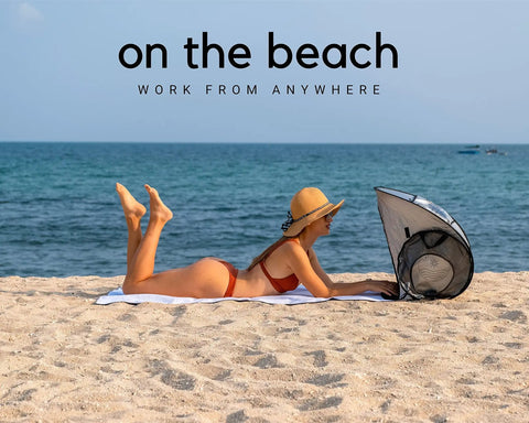Woman using laptop under Outdoor Laptop Sun Shade For Working Anywhere Glare Blocker And Heat Reflective Hood on the beach.