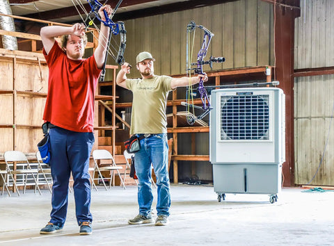 Two men with bows practicing archery in a barn with a Heavy Duty Evaporative Air Cooler With Killer Cooling Power nearby.