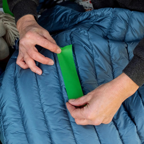 Applying Tenacious Tape 3 Inch By 20 Inch Clear Vinyl Gear Repair Tape to a blue sleeping bag for quick hole and tear fixes.