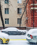 Cars parked on a snowy street with snow-covered trees and buildings in the background during winter snowfall.