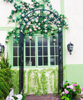 Sturdy Black Wrought Iron Pergola Arbor Garden Arch With Ground Spikes 100 Inches High And 45 Inches Wide Outdoor Climbing Plants Trellis For Weddings Parties And Patio Decor adorned with flowers against a building.