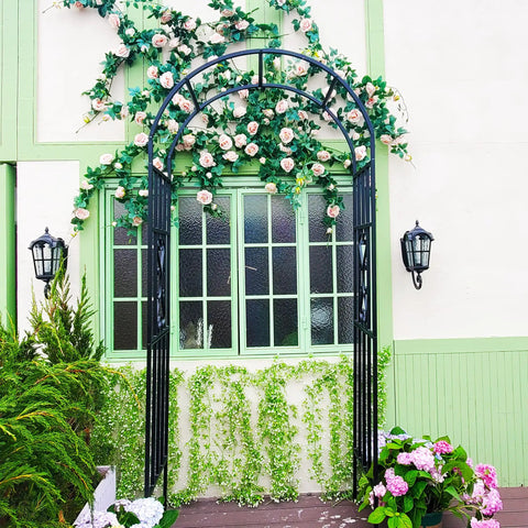 Sturdy Black Wrought Iron Pergola Arbor Garden Arch With Ground Spikes 100 Inches High And 45 Inches Wide Outdoor Climbing Plants Trellis For Weddings Parties And Patio Decor adorned with flowers against a building.