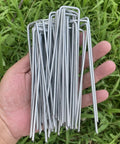 Hand holding sturdy 6 inch galvanized steel garden stakes for securing weed fabric landscape netting ground sheets fleece