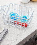 White Coated Steel Cabinet Organizer Basket With Snap Lid holding pacifiers and utensils, durable kitchen storage on countertop.