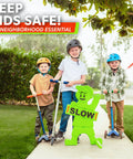Kids at Play Safety Sign with high-visibility flag on sidewalk with children on scooters for traffic slow down zone