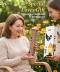 Two women exchanging a boxed bird feeder gift beside chair and flowers, product packaging is visible