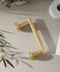 Gold 2 Pack Pivoting Toilet Paper Holder In SUS304 Stainless Steel Wall Mounted Double Post Tissue Roll Holder For Commercial Bathrooms And RVs Rustproof And Built To Last shown on a wooden surface with a roll of toilet paper nearby and decorative greenery.