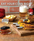 Grilled corn with metal holders on a wooden board with BBQ brush beside, beer and garlic scene.