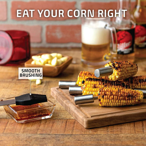 Grilled corn with metal holders on a wooden board with BBQ brush beside, beer and garlic scene.
