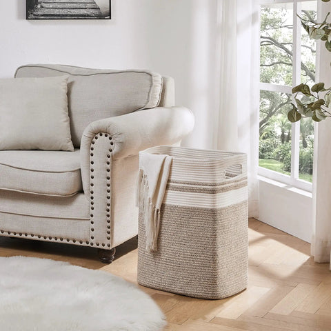 Beige storage basket by a sofa in a living room with a blanket draped over, showcasing All In One Cotton Rope Laundry Hamper And Storage Basket With Handles Collapsible Go To Organizer For Living Room Toys Blankets And Clothes Yellow Variegated 16 By 13 By 22 Inches.