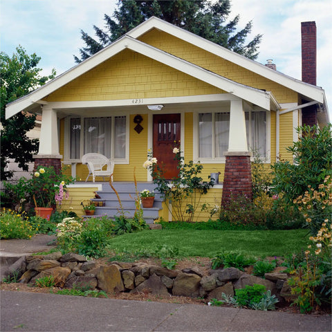 Charming yellow bungalow with a spacious porch surrounded by greenery and a stone-bordered front garden.