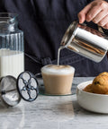 Handheld Milk Frother For Creamy Foam In Seconds, On The Go Stainless Steel Foamer in use with cup of coffee on table