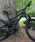 Mountain bike with Rear Derailleur Guard For Mountain Bikes And EBikes, Ultra Lightweight And Easy To Install, Universal Fit No Modifications Needed, Protect Your Derailleur From Rocks, Roots And Obstacles On The Trail Made In USA, leaning against a tree