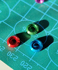 Colorful aluminum M3 counterbore washers in green, red, and blue on a drafting mat, part of Top Shelf 60 Pack M3 Counterbore Washers In Sweet Golden Anodized Aluminum Alloy Finish And Rock Solid Grip Perfect For Rc Drone Fpv Toys Mechanical Screw Sets No Sweat Assembly.