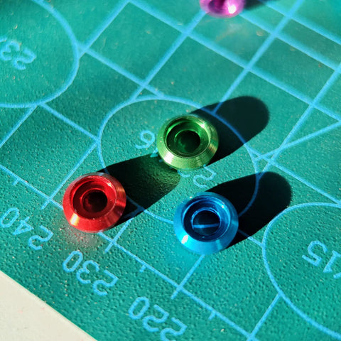 Colorful aluminum M3 counterbore washers in green, red, and blue on a drafting mat, part of Top Shelf 60 Pack M3 Counterbore Washers In Sweet Golden Anodized Aluminum Alloy Finish And Rock Solid Grip Perfect For Rc Drone Fpv Toys Mechanical Screw Sets No Sweat Assembly.