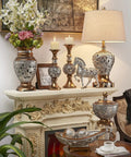 Langi Table Lamp With Mirrored Tile Neck Bronze Base With Silver Leaf Floral Accents Beige Drum Shade On Mantel