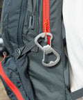 Dual Bottle Opener And SBiner Carabiner Combo In Stainless Steel attached to a backpack near a red zipper.