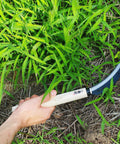 Hand holding Go-To 6.7 Inch Japanese Serrated Garden Sickle with razor sharp carbon steel blade, cutting through weeds in garden.
