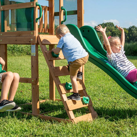 Child sliding down green wave slide on Buckley Hill Cedar Wood Swing Set With Wave Slide, Elevated Play Deck, Rock Wall Ladder, Belt Swings.