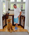 Man standing with dog behind 4-Panel Freestanding Pet Gate With Adjustable Wooden Panels For Doorways, Ideal For Dogs And Cats, 36 Inches Tall By 76 Inches Wide, Walnut Finish, Ready To Use Right Out Of The Box.