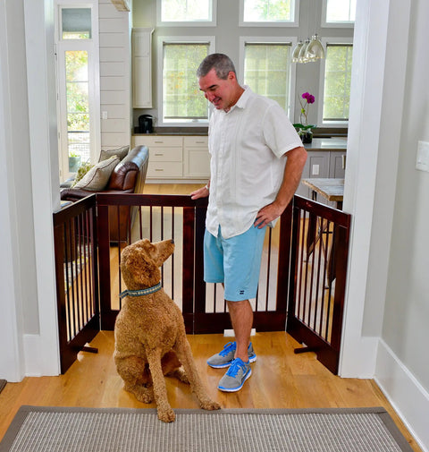 Man standing with dog behind 4-Panel Freestanding Pet Gate With Adjustable Wooden Panels For Doorways, Ideal For Dogs And Cats, 36 Inches Tall By 76 Inches Wide, Walnut Finish, Ready To Use Right Out Of The Box.