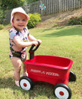 Child using a red push and pull walker wagon with garden tools for kids one to four years old in a grassy yard.