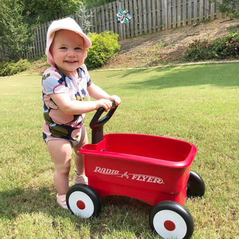 Child using a red push and pull walker wagon with garden tools for kids one to four years old in a grassy yard.