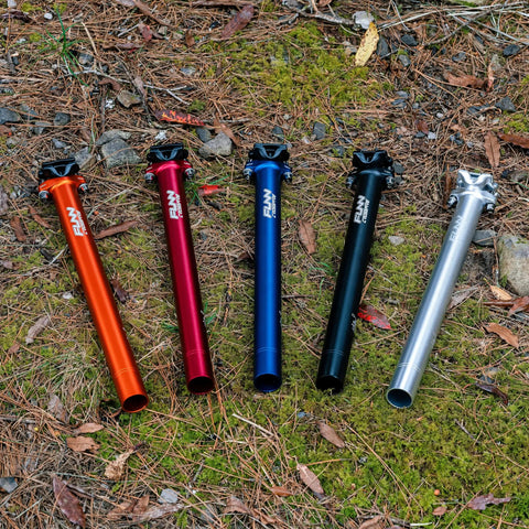 Colorful bike seat posts in various shades including orange, red, blue, black, and silver displayed on forest ground.