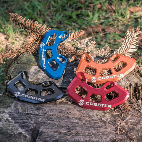 Four colorful Cogster bash guards on a log, including black FortiGuard Alloy MTB Bash Guard ISCG05 Compatible For 26T To 36T Chainrings.