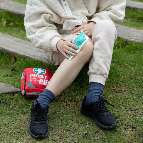 Red compact first aid kit bag on grass; person holding a blue instant cold pack.