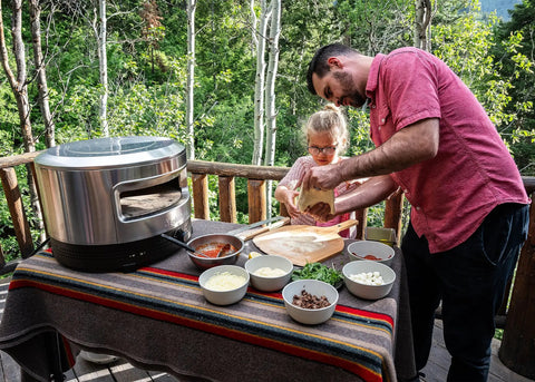 Father and daughter using Outdoor Pizza Oven On The Go For Gas Or Propane Cooking with toppings on a table outdoors