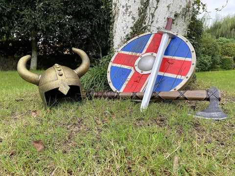 Kids Viking Horned Helmet For Costume Play LARP And Halloween on grass with shield, sword, and axe on a tree trunk.