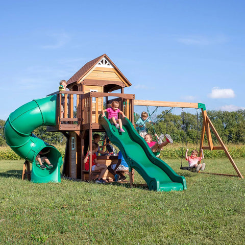 Children playing on Cedar Cove Wooden Swing Set With Spiral Tube Slide And Wave Slide, Elevated Clubhouse, and Swings in backyard.